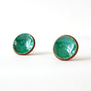 Puede incluir: Un par de pendientes de cerámica verde con un borde marrón. Los pendientes son redondos y tienen una forma ligeramente abovedada.