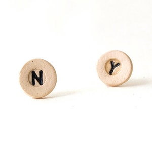 Puede incluir: Un par de pendientes de cerámica con las letras "N" e "Y" pintadas en negro sobre un fondo beige.
