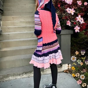 May include: A woman wearing a pink, blue, and purple hooded sweater dress with black leggings and black sneakers. The dress has a layered ruffle hem and a colorful patterned design. The woman is standing on a set of stairs with a flower bush in the background.