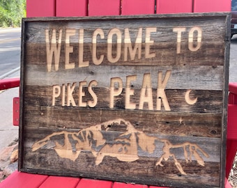 May include: Wooden sign with the words "WELCOME TO PIKES PEAK" carved into the wood. The sign also features a mountain range silhouette and a crescent moon. The sign is made of reclaimed wood and has a rustic appearance.