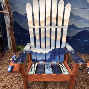 May include: A wooden chair made from repurposed skis. The chair has a brown wooden frame and a blue and white painted ski design. The chair is sitting on a bed of brown mulch.