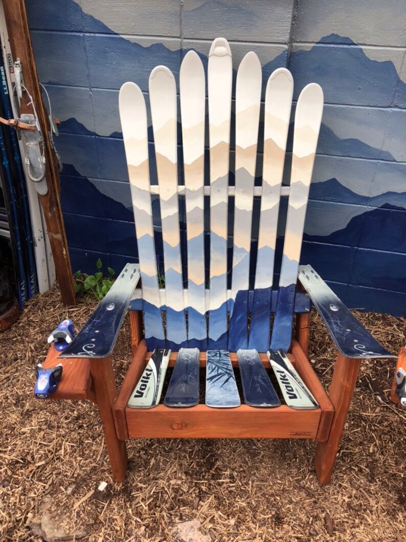 May include: A wooden chair made from repurposed skis. The chair has a brown wooden frame and a blue and white painted ski design. The chair is sitting on a bed of brown mulch.