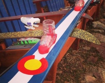 May include: A blue and white ski with a red, yellow, and white circle painted on it. The ski is propped up on a wooden surface and has several clear glass bottles attached to it.
