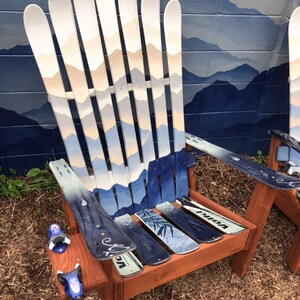 May include: A wooden chair made from repurposed skis. The chair is painted with a mountain scene in shades of blue and white. The chair has a wooden seat and back, and the skis are used as the chair's legs and back support. The chair is sitting on a bed of brown straw.
