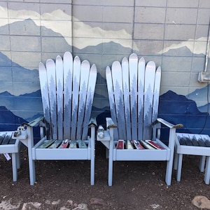 May include: Two gray Adirondack chairs made from repurposed skis, with a mountain mural painted on the wall behind them. The chairs have a gray wooden frame and a seat made from skis. There are two small gray wooden stools in front of the chairs.