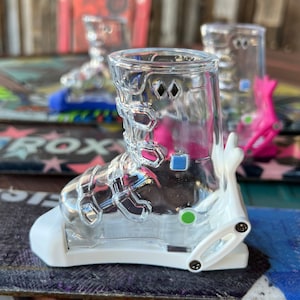 May include: Clear glass shot glass in the shape of a ski boot. The boot has a white base and accents, with a green and blue dot. The shot glass is sitting on a snowboard.