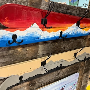 May include: Hand-painted wooden coat racks with black metal hooks. The top rack is red with a sunset and mountain scene. The middle rack is blue with a mountain scene. The bottom rack is beige with a mountain scene.