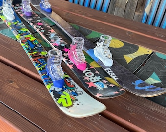 May include: Four pairs of skis with colorful designs and shot glasses attached to them. The skis are on a wooden table.