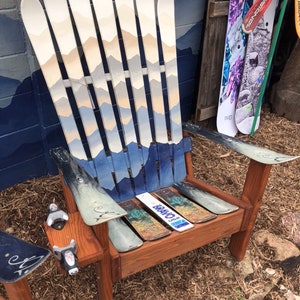 May include: A wooden chair made from repurposed skis. The chair has a mountain scene painted on the back and the seat is made from a wooden plank. The chair is sitting on a bed of brown straw.