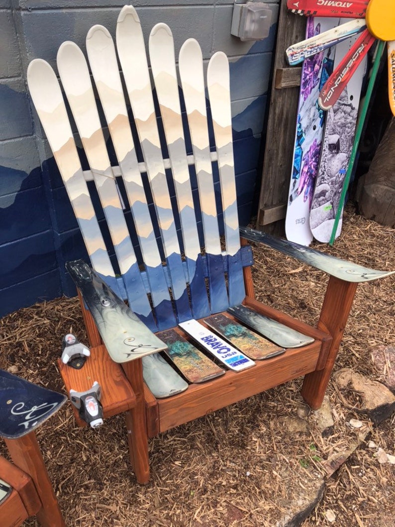 May include: A wooden chair made from repurposed skis. The chair has a mountain scene painted on the back and the seat is made from a wooden plank. The chair is sitting on a bed of brown straw.