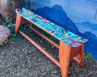 May include: A wooden bench made from skis. The skis are decorated with colorful graphics and are mounted on a wooden frame. The bench is set against a blue wall with a mountain mural.