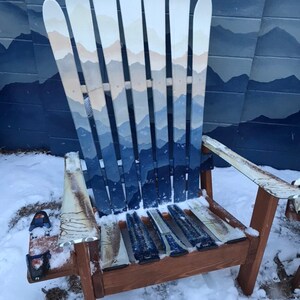 May include: A wooden chair made from painted skis. The chair is painted with a blue and white mountain scene. The chair is sitting in the snow.