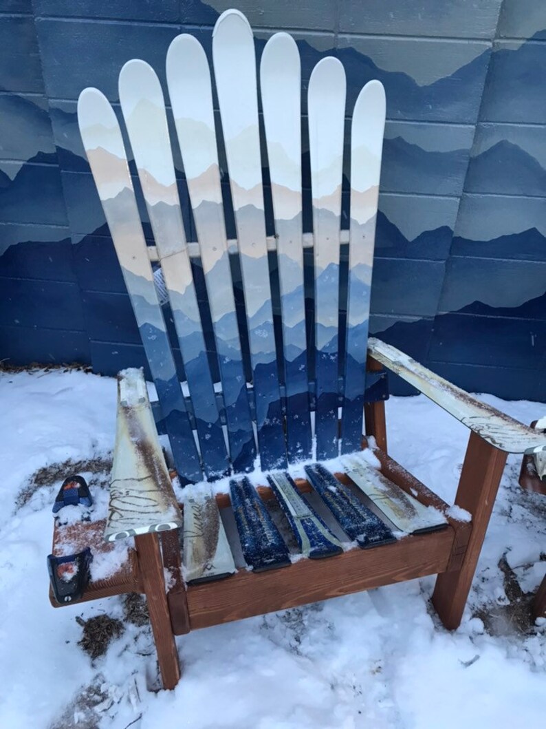 May include: A wooden chair made from painted skis. The chair is painted with a blue and white mountain scene. The chair is sitting in the snow.
