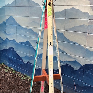 May include: A wooden ski rack with three pairs of skis. The skis are white with pink and blue accents. The rack is against a blue and white brick wall with a mountain mural.