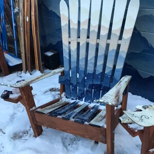 May include: A wooden chair made from multiple blue and white painted skis. The chair is sitting in the snow. The chair has a mountain scene painted on the back.