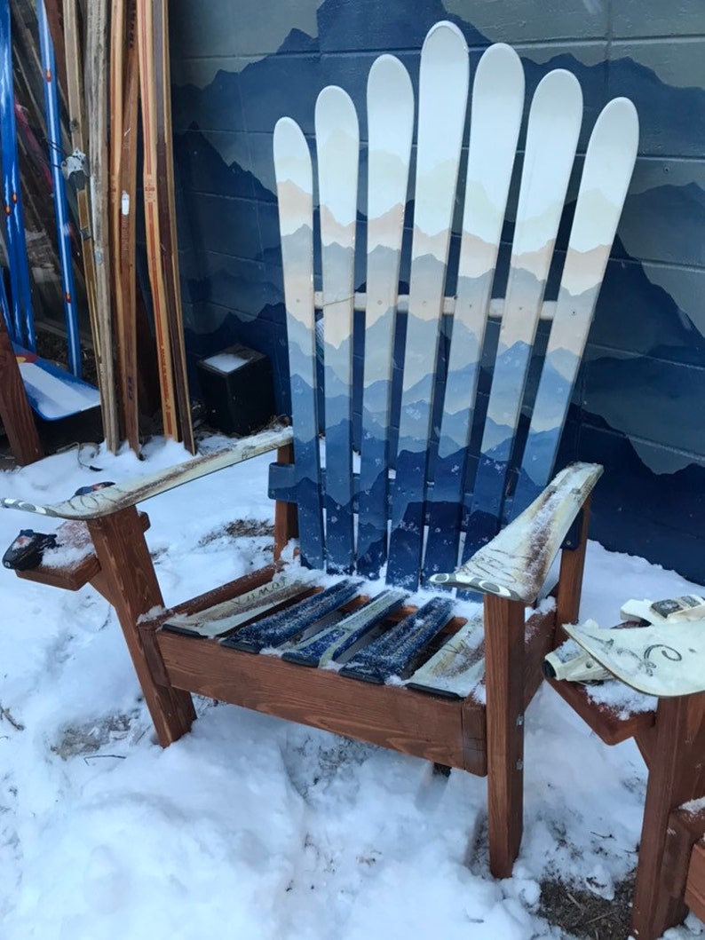 May include: A wooden chair made from multiple blue and white painted skis. The chair is sitting in the snow. The chair has a mountain scene painted on the back.