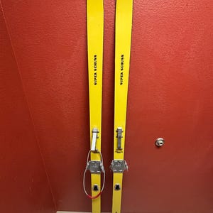 May include: A pair of vintage yellow skis with black lettering that reads "SUPER SCHUSS" on each ski. The skis have metal bindings and are in good condition.