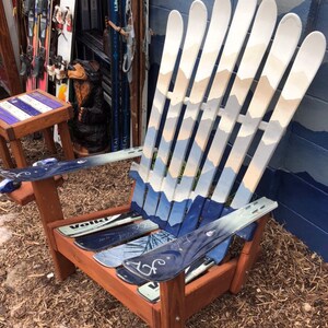May include: A wooden Adirondack chair made from repurposed skis. The chair is painted white with blue and beige mountain scenes. The chair is sitting on a bed of brown mulch.