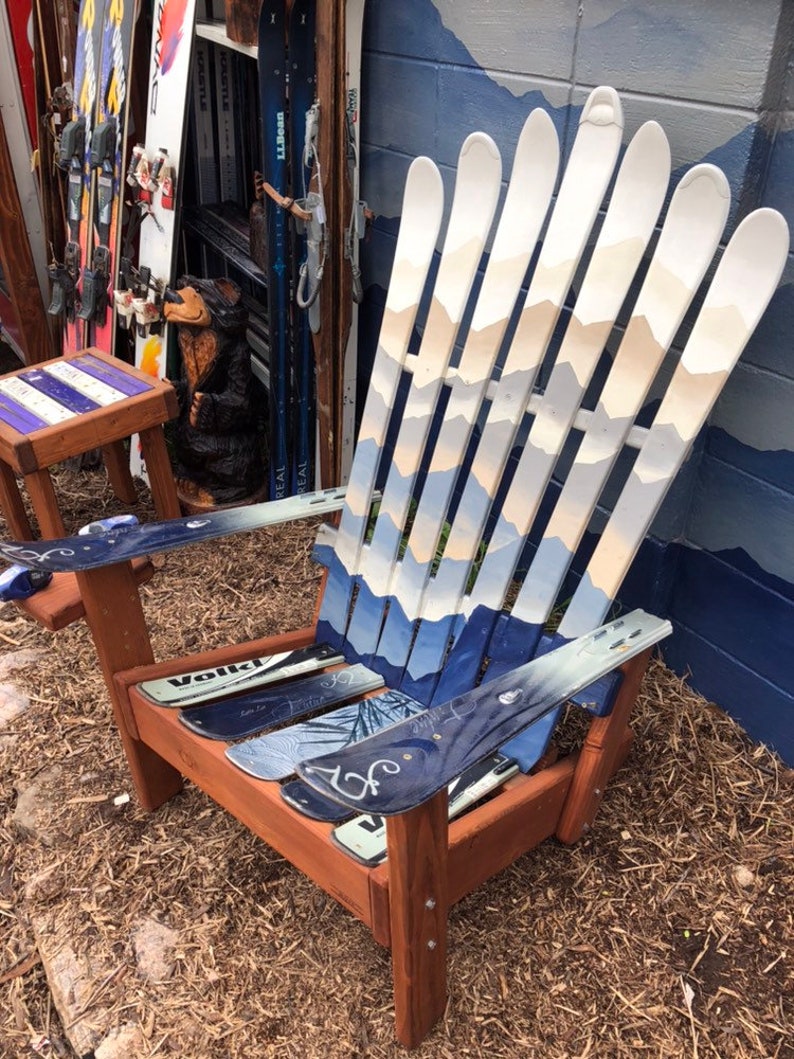 May include: A wooden Adirondack chair made from repurposed skis. The chair is painted white with blue and beige mountain scenes. The chair is sitting on a bed of brown mulch.