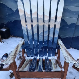 May include: A wooden chair made from painted skis. The chair is painted with a mountain scene in shades of blue and white. The chair is sitting in the snow.