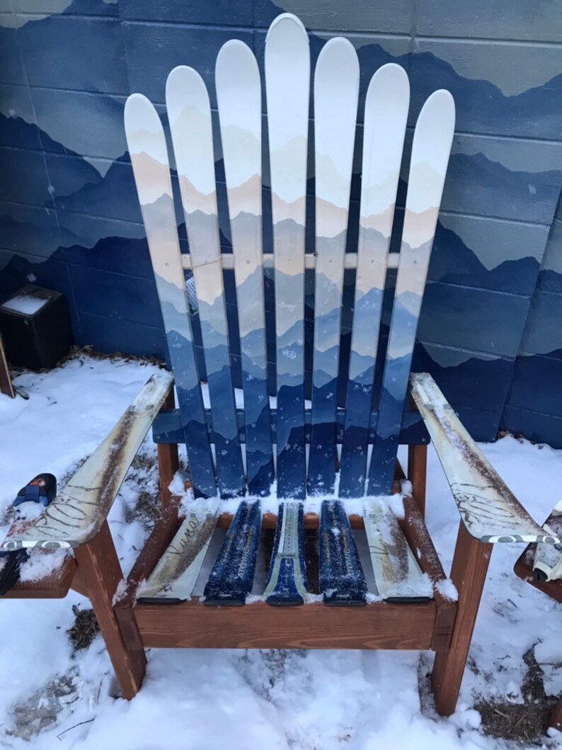May include: A wooden chair made from painted skis. The chair is painted with a mountain scene in shades of blue and white. The chair is sitting in the snow.