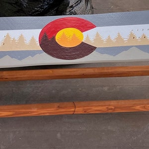 May include: A wooden bench with a painted snowboard top. The snowboard is painted with the Colorado state flag, featuring a yellow sun, red and white stripes, and a brown mountain range. The bench is made of natural wood with a reddish-brown stain.