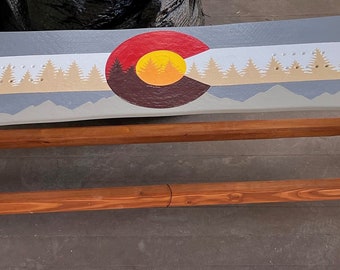 May include: A wooden bench with a painted snowboard top. The snowboard is painted with the Colorado state flag, featuring a yellow sun, red and white stripes, and a brown mountain range. The bench is made of natural wood with a reddish-brown stain.