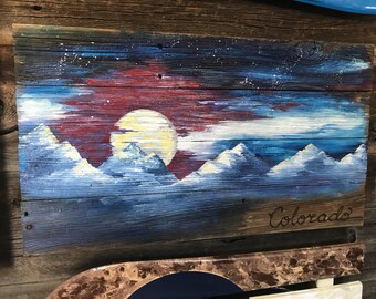 May include: A wooden sign with a painted scene of a mountain range at night with a full moon and stars. The sign is painted in shades of blue, red, white, and yellow. The words "Colorado" are written in black on the bottom of the sign.