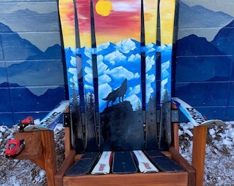 May include: A wooden chair made from repurposed skis, painted with a sunset scene, mountains, and a wolf howling at the moon. The chair has a wooden armrest and a ski for a footrest.