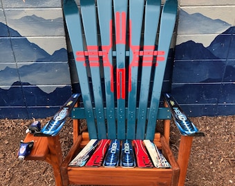 May include: A teal blue Adirondack chair made from repurposed skis. The chair has a wooden frame and a ski-shaped back. The seat is made of skis with red, white, and blue designs. The chair is sitting on a bed of brown wood chips.