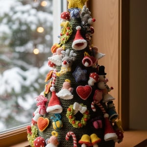 Op de afbeelding: Een handgemaakte, gehaakte kerstboom met diverse ornamenten. De boom is donkergroen en versierd met rode, witte en gele gehaakte decoraties, waaronder harten, sterren en kerstmutsen. Hij staat op een houten vensterbank.