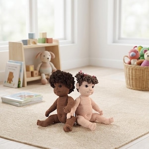 Op de afbeelding: Twee handgemaakte gehaakte babypoppen, één bruin en één roze, zitten op een beige tapijt. De poppen hebben krullend haar en kijken naar voren. Op de achtergrond een houten boekenplank met speelgoed en boeken, samen met een rieten mand vol knuffels.