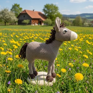 Op de afbeelding: Een gehaakte ezel figuur in een veld met gele paardenbloemen. De ezel is grijs met bruine accenten op zijn manen, staart en hoeven. Hij staat op een kleine, ronde, beige basis. Een huis met een rood dak is op de achtergrond.