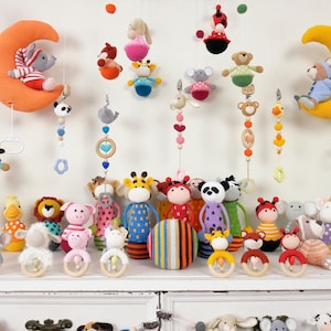 Op de afbeelding: Een verzameling handgemaakte gehaakte baby mobielen en speelgoed. De afbeelding toont verschillende mobielen met dierenthema's, waaronder beren, giraffen en olifanten, samen met bijpassende rammelaars. Het kleurenpalet omvat oranje, geel, blauw en roze.