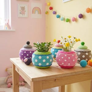 Op de afbeelding: Een collectie ronde, gehaakte hoezen voor potten, in levendige kleuren zoals paars, limoengroen, blauw en roze, allemaal versierd met witte stippen. Sommige hoezen hebben bijpassende deksels, terwijl andere een vetplant of madeliefjes bevatten.