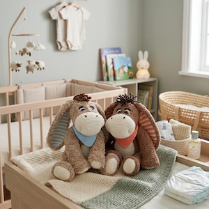 Op de afbeelding: Twee gehaakte ezels met grote oren en bandana's zitten op een aankleedtafel. De ezels zijn bruin en beige met blauwe en oranje bandana's. Een gebreide deken met groene en beige strepen ligt eronder. De kinderkamer heeft een wieg en een mobiel.