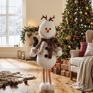 May include: A festive snowman ornament with a fluffy white body, brown scarf, and carrot nose. It features twig antlers and stands on a wooden base. The snowman is in a room with a decorated Christmas tree and a comfortable armchair.