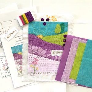 Könnte beinhalten: Ein DIY-Stickset mit einem farbenfrohen Landschaftsdesign. Das Set enthält einen vorgeprägten Stoff, Stickgarn und Stoffreste in den Farben Lila, Grün und Blau. Das Set enthält auch Anleitungen zum Sticken des Designs. Das Set trägt den Titel "Slow Stitch by Fairy Floo 1" und enthält den Text "Be Positive".