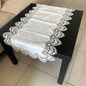 May include: A black rectangular coffee table with a white lace table runner. The runner features a floral pattern and a scalloped edge. The table is made of wood and has four legs.