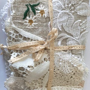 Puede incluir: Un conjunto de retales de tela en varios tonos crema y beige, atados con una cinta marr&oacute;n claro. La pieza superior presenta flores blancas bordadas con hojas verdes. Se aprecian detalles de encaje y ganchillo.