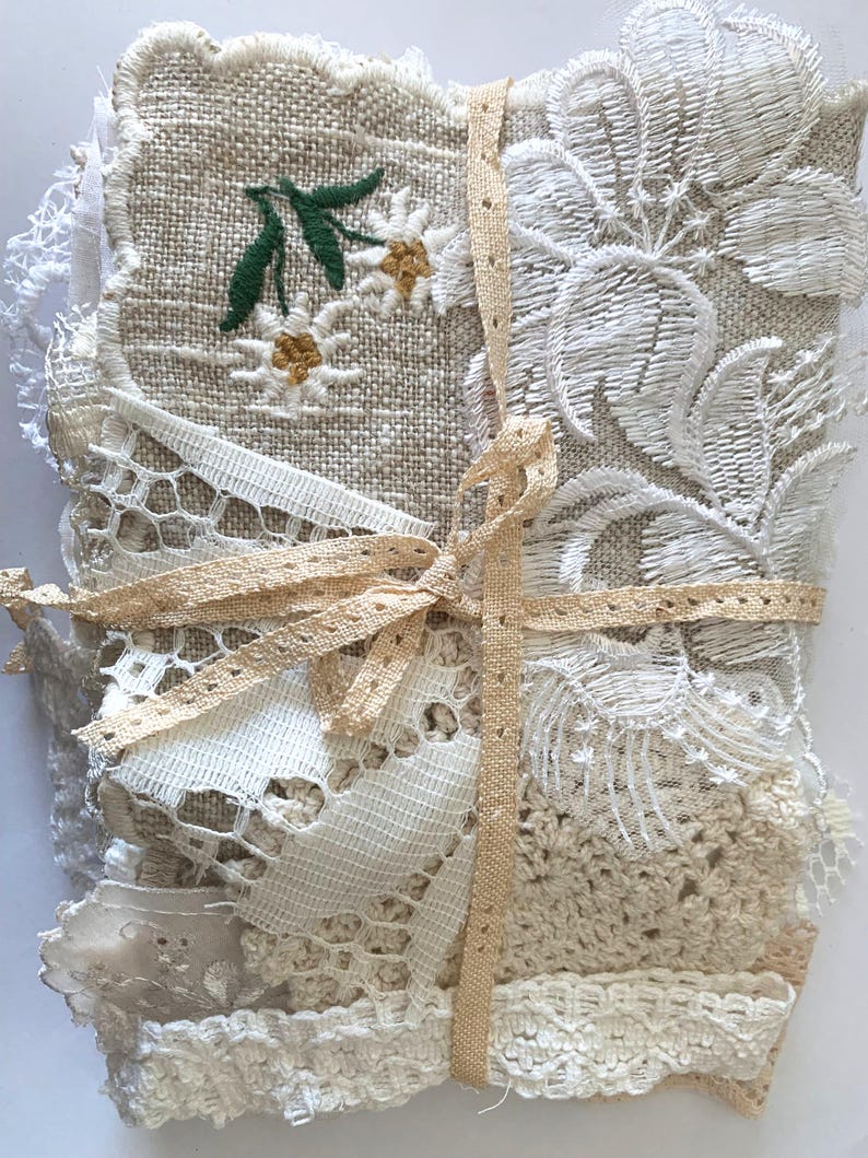 Puede incluir: Un conjunto de retales de tela en varios tonos crema y beige, atados con una cinta marr&oacute;n claro. La pieza superior presenta flores blancas bordadas con hojas verdes. Se aprecian detalles de encaje y ganchillo.