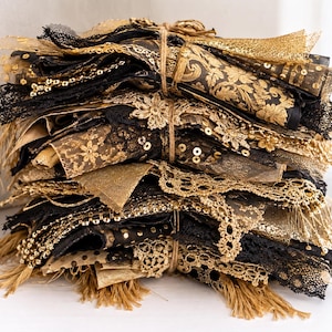 Könnte beinhalten: Ein Stapel verschiedener Stoffreste in Schwarz- und Goldtönen, die mit einer Schnur zusammengebunden sind. Die Sammlung umfasst verschiedene Texturen wie Spitze, Paillettennetz, florales Brokat und Fransenränder, die ein geschichtetes Bündel bilden.