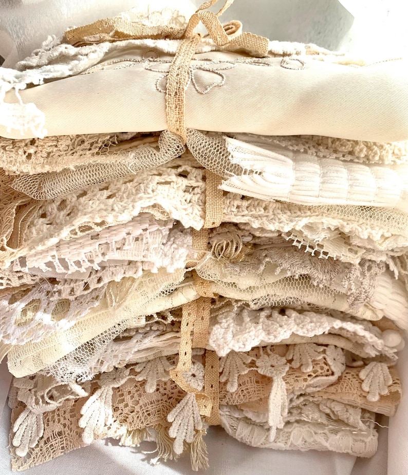 Puede incluir: Una pila de encajes y telas vintage en varios tonos crema y blanco roto, atada con una cinta beige. Los textiles incluyen patrones de ganchillo, bordados y encajes delicados. Las texturas y los colores sugieren una colecci&oacute;n de ropa de cama antigua o suministros de artesan&iacute;a.