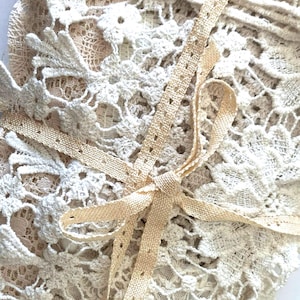 Puede incluir: Primer plano de tela de encaje color crema con intrincados patrones florales. Una delicada cinta beige est&aacute; atada en un lazo, acentuando la textura y el detalle del encaje. La imagen resalta la artesan&iacute;a del textil.