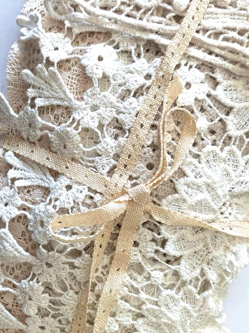 Puede incluir: Primer plano de tela de encaje color crema con intrincados patrones florales. Una delicada cinta beige est&aacute; atada en un lazo, acentuando la textura y el detalle del encaje. La imagen resalta la artesan&iacute;a del textil.