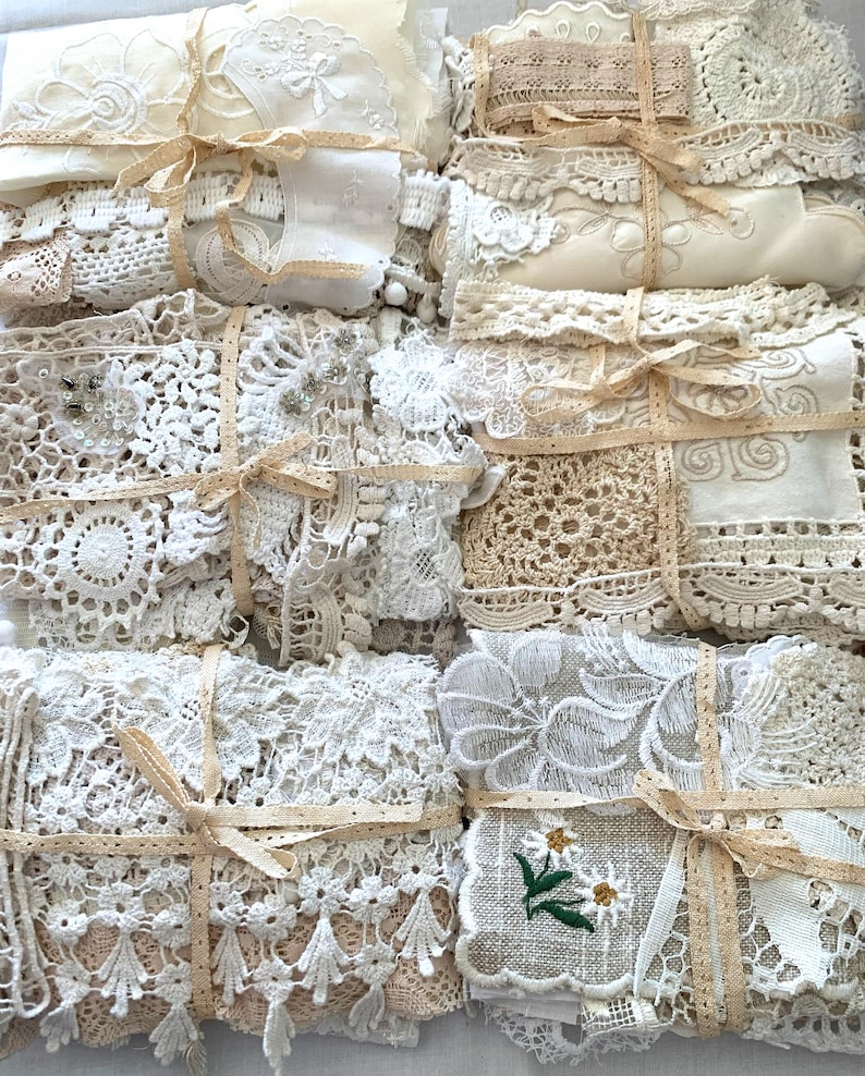Puede incluir: Un surtido de paquetes de encaje y tela vintage atados con cintas beige. Los paquetes presentan varias texturas, incluyendo ganchillo, dise&ntilde;os florales bordados y delicados adornos de encaje. La paleta de colores es principalmente crema y beige.