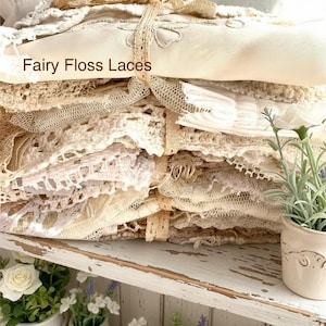 Puede incluir: Una pila de textiles de encaje color marfil y crema, atados con una cinta beige. El texto "Fairy Floss Laces" es visible. El encaje presenta diversos patrones y texturas. La pila est&aacute; sobre una estanter&iacute;a blanca con peque&ntilde;as plantas en macetas.