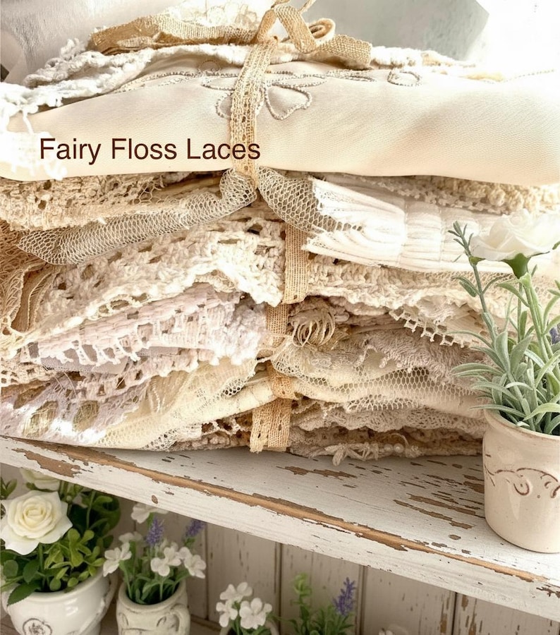 Puede incluir: Una pila de textiles de encaje color marfil y crema, atados con una cinta beige. El texto "Fairy Floss Laces" es visible. El encaje presenta diversos patrones y texturas. La pila est&aacute; sobre una estanter&iacute;a blanca con peque&ntilde;as plantas en macetas.