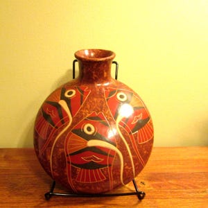 May include: A red, brown, and black ceramic vase with a fish design. The vase is on a wooden table.
