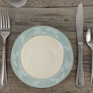 May include: A light blue and white patterned ceramic plate with a white rim. The plate is empty and is set on a wooden table with a silver fork, knife, and spoon beside it.